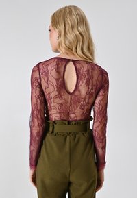 Body in pizzo borgogna con maniche lunghe e apertura a chiave sul retro, abbinato a pantaloni olive verdi a vita alta con vita arricciata.