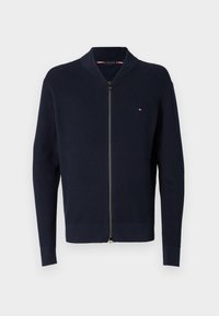 Cardigan tricotat navy cu închidere cu fermoar, textură în relief și manșete contrastante. Prezintă un mic logo pe partea stângă a pieptului.
