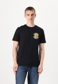 Svart kortärmad t-shirt med rundhals, prydd med en gul solrosgrafik och texten "SUNNIE'S POOL BAR." Bärs med blå jeans.