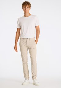 Beige chinos med slim fit, parret med en ensfarvet hvid crew neck t-shirt. Modellen har hvide sneakers på og står foran en ensfarvet baggrund.