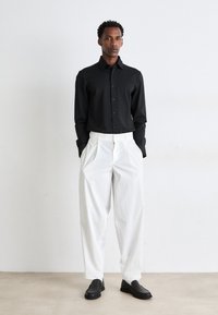 Camisa negra de botón con tela texturada, combinada con pantalones blancos de corte holgado y zapatos negros sin cordones sobre un fondo neutro.