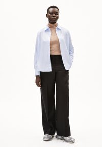 Lichtblauw button-up shirt over een beige coltrui, gecombineerd met zwarte wijde broek en grijze sneakers, op een neutrale achtergrond.