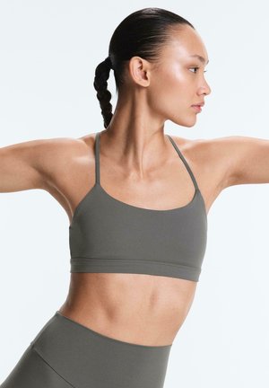 Kvinde med flettet hår iført grå sports-bh og leggings, der strækker armene ud til siderne mod en ensfarvet hvid baggrund.