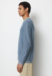 Blau gestreifter Strickpullover mit langen Ärmeln, geripptem Ausschnitt und Bündchen sowie angenehmer Textur. Über hellen Hosen getragen.
