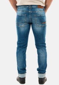 Jeans azules deslavados con un ajuste slim, que cuentan con un parche de cuero, bolsillos traseros con detalles de costura y puños doblados sobre zapatos negros.