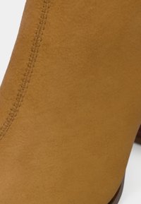 Közelkép egy világosbarna színű, visszavarrt varrással ellátott szarvasbőr cipőről, amelynek része egy alacsony barna sarok is látható fehér háttéren.