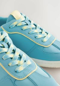 Ljusblå sneakers med gula detaljer, texturerade blå tygskosnören, vadderad plös och sömnadsdetaljer. Vit gummisul.