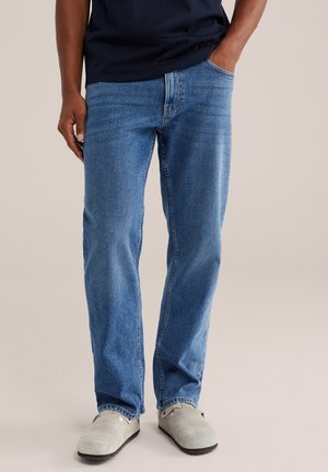 Mann trägt blaue Jeans mit geradem Schnitt, eine Hand in der Tasche und hellgraue Slipper, steht vor einem schlichten Hintergrund.
