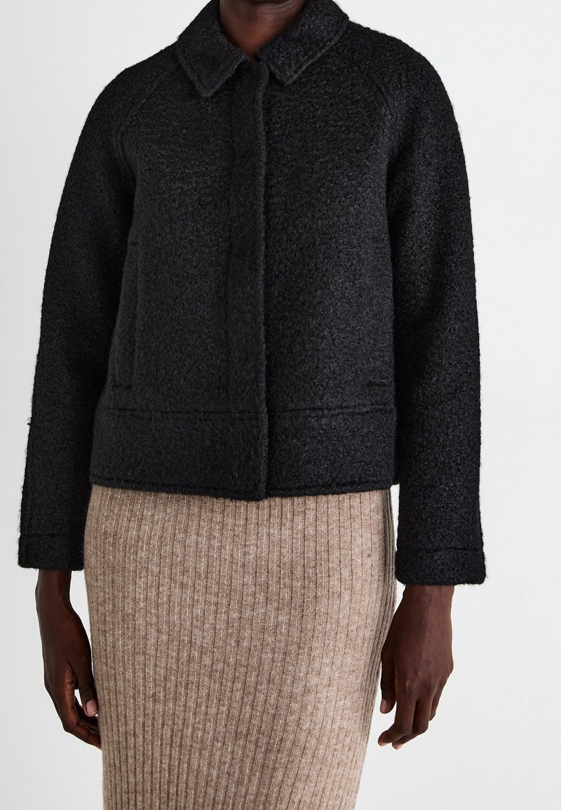 Personne portant une veste en laine texturée noire avec col et manches longues, associée à une jupe en maille côtelée beige.