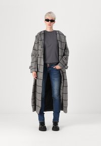 Person with short blond hair wearing black sunglasses, gray checkered long coat, dark gray shirt, blue jeans, and black boots, standing.