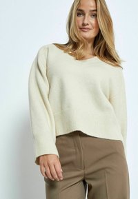 Maglione color crema con scollo a V, orlo e polsini a costine; texture morbida, lunghezza leggermente cropped, indossato su pantaloni sartoriali marrone chiaro.