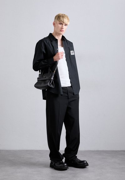Jeune personne avec des cheveux blonds courts, portant un t-shirt noir avec un patch de code-barres, un débardeur blanc, un pantalon noir et des chaussures noires imposantes, tenant un sac à main noir.