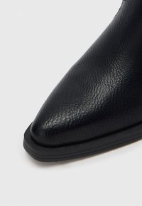 Čierna členková topánka z umelej kože s špicatou špičkou, textúrovaným povrchom a plochou čiernou podrážkou s jemnými horizontálnymi líniami.