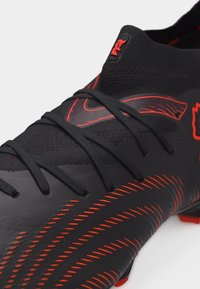 Μαύρο και κόκκινο ποδοσφαιρικό παπούτσι με επάνω μέρος από πλεκτό υλικό, κορδόνια, υφή στην σόλα και λεπτομερείς ραφές κατά μήκος της πλευράς και της κορυφής.