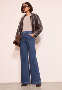 Rjava usnjena jakna čez bež zgornji del, kombinirana z visokim pasom in širokimi modrimi kavbojkami. Dodatki vključujejo rjavo torbico in sončna očala.