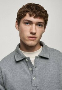 Grå pikétröja med krage, med en knappslå, som bärs över en ljusfärgad t-shirt med rund hals. Tyget verkar mjukt och något texturerat.