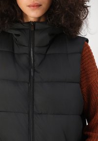 Veste sans manches noire matelassée avec fermeture éclair à l'avant et capuche, présentant une texture matelassée. Portée par-dessus un pull en tricot couleur rouille.