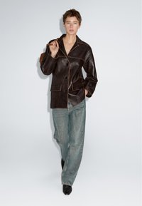 Persoon met kort haar, draagt een donkerbruine leren jack en loszittende blauwe jeans, met een bruine tas over één schouder, die naar voren loopt.