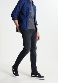 Jeans oscuros combinados con una camisa gris y una camisa de cuadros azules, complementados con zapatillas negras con detalles blancos. Un atuendo casual con estilo en capas.