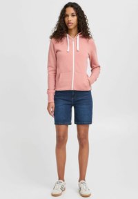 Rosa zip-up hoodie med vita dragsnören, framfickor och långa ärmar, ihop med mörka denimshorts och vita sneakers.