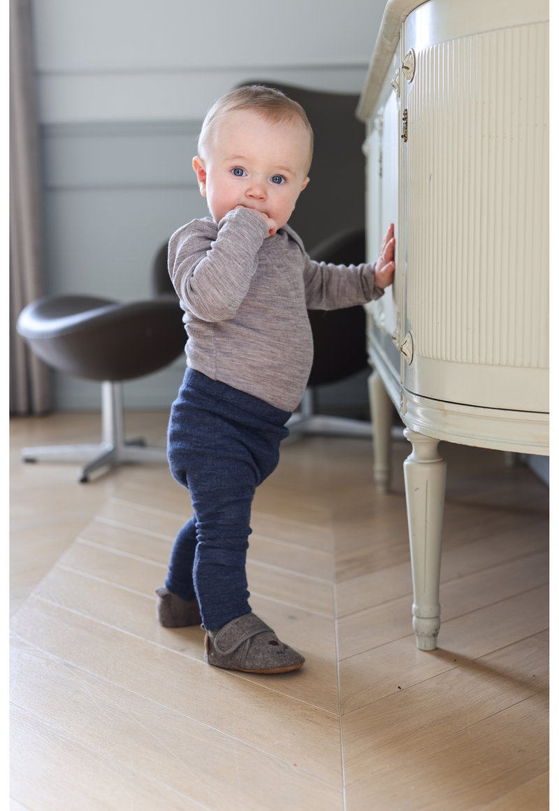 Enfant portant un t-shirt à manches longues gris et des leggings bleu marine, debout à côté d'un meuble crème avec des rainures verticales ; chaussures grises en feutre.