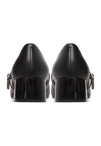 Een paar zwarte leren schoenen met lage blokhak, enkelbandjes en metalen gespen, gezien van achteren tegen een witte achtergrond.