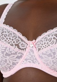Zbliżenie na ciemną skórę ubraną w jasno-różowy koronkowy biustonosz z kwiatowymi wzorami oraz małą kokardką i zawieszką na środku.