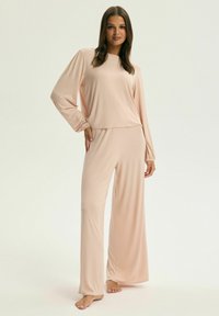 Jasnoróżowy zestaw wypoczynkowy, składający się z bluzki z długim rękawem, wyposażonej w elastyczne mankiety, oraz spodni z szerokimi nogawkami. Miękka tkanina o gładkiej fakturze i luźnym kroju.