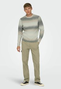 Lys grå sweater med mørkegrå og beige gradientstriber, ribbede manchetter, parret med olivengrønne bukser og sorte canvas sneakers.