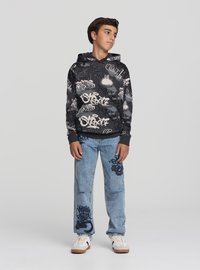 Felpa nera con testo in stile graffiti bianco, abbinata a jeans azzurri strappati con motivi grafici simili e sneakers bianche.