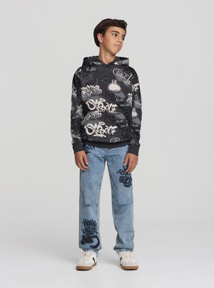 Felpa nera con testo in stile graffiti bianco, abbinata a jeans azzurri strappati con motivi grafici simili e sneakers bianche.