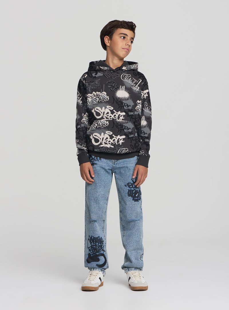 Felpa nera con testo in stile graffiti bianco, abbinata a jeans azzurri strappati con motivi grafici simili e sneakers bianche.