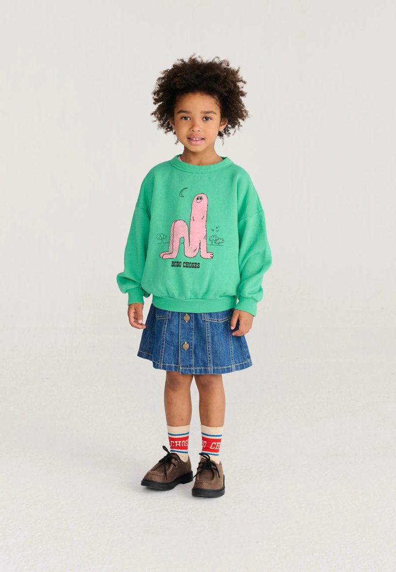 Bambino piccolo con capelli ricci che indossa una felpa verde con un cartone animato rosa, gonna di jeans blu, calzini a righe e scarpe marroni, in piedi su uno sfondo semplice.