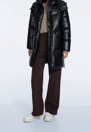 Personne portant une longue doudoune noire, un pantalon large à rayures marron, un haut beige et des chaussures blanches épaisses sur un fond uni.