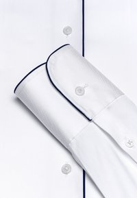 Weißes Hemd mit glatter Textur, marineblauer Paspel entlang des Kragens und der Manschetten sowie transparenten Knöpfen. Die Manschetten sind aufgerollt und ordentlich gefaltet.