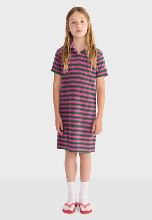 Mädchen steht und trägt ein kurzärmeliges Kleid mit pink-grünen Streifen, weiße Socken und rote Flip-Flops vor einem einfarbigen Hintergrund.