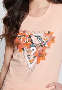Pfirsichfarbenes ärmelloses T-Shirt mit einem floralen Dreiecks-Design, das "GUESS" in Weiß, funkelnde Akzente und filigrane Design-Details zeigt.