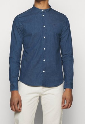 Mężczyzna w dopasowanej, ciemnoniebieskiej koszuli zapinanej na guziki z kołnierzykiem typu stójka i białych spodniach, stojący na jednolitym tle.