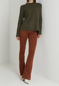 Person in olivgrünem geripptem Pullover und rostfarbenen Schlaghosen mit weißen Absatzstiefeln, stehend vor einer schlichten hellen Wand.