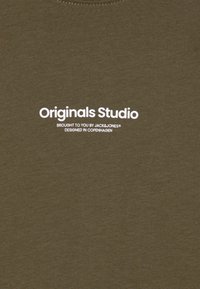 Olive spalvos medvilnės marškinėliai su baltais spausdintais užrašais, kuriuose užrašyta „Originals Studio“, ir prekės ženklo informacija žemiau.
