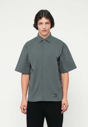 Kortærmet skjorte med knapper i mørkegrå, med en struktureret krave, subtil tekstur og sidelommer med lynlås og et label-detalje.