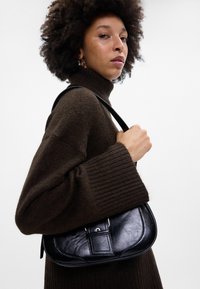 Melnas ādas rokassomu ar izliekta formu, ar atloka aizdari un regulējamu siksnu, tur rokās pret tumšu džemperi neitrālā fona priekšā.