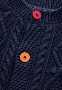 Tengerészkék kötött szövet vastag kötélmintákkal és két színes, "Little Bird" feliratú gombbal, egy piros és egy narancssárga, varrva.