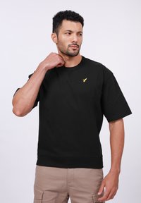 T-shirt en coton noir avec des manches courtes, un col rond et un petit logo doré sur la poitrine. Tissu texturé, coupe décontractée.