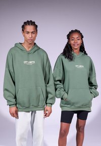 Grønne overdrevne hettegensere med en frontlomme og "OFF LIMITS" trykket i hvitt. Uformell passform, brukt med lyse bukser og sorte shorts.
