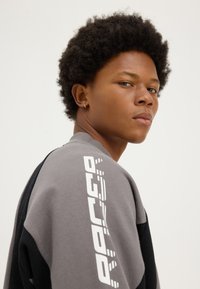 Jeune personne aux cheveux courts et bouclés, portant une petite boucle d'oreille en or, vêtue d'un sweat-shirt gris et noir avec un texte blanc sur la manche.
