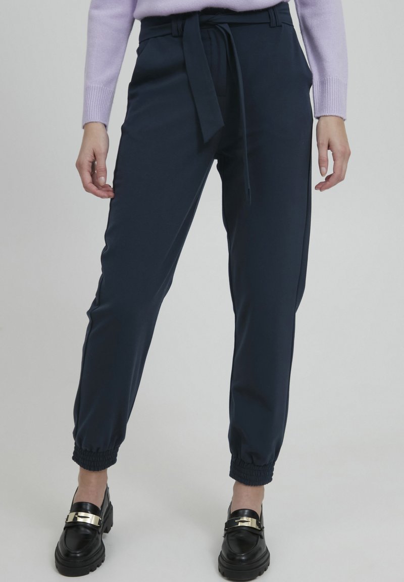 Navy blue, high-waisted trousers with a belted waist, tapered ankles, and elastic cuffs, paired with black shoes featuring a gold accent.