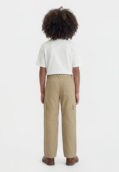 Niño con cabello rizado de espaldas, vestido con una camiseta blanca, pantalones cargo beige y zapatos marrones sobre un fondo liso.