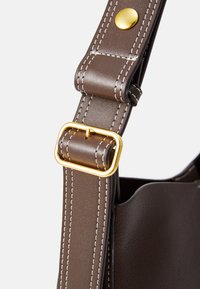 Bandoulière en cuir marron avec des coutures blanches, comportant une boucle en métal doré et un bouton pression. Design avec des bords arrondis visible.