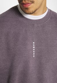 Felpa di cotone viola con collo alto, polsini a costine e stampa verticale bianca con la scritta "NOWHERE" sul davanti.